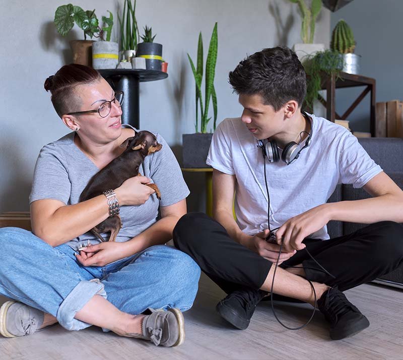 Happy talking mom and teenage son, parent and child sitting at home on the floor with pet little dog toy terrier, real friendly loving family