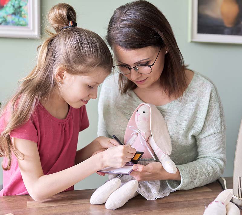 Mother and daughter child play together at home, girl and mom make make-up paints cheeks to toy handmade doll bunny sewn on sewing machine